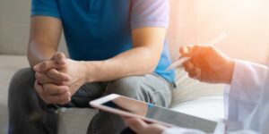 Patient sitting on exam table with hands crossed while doctor holds tablet and talks